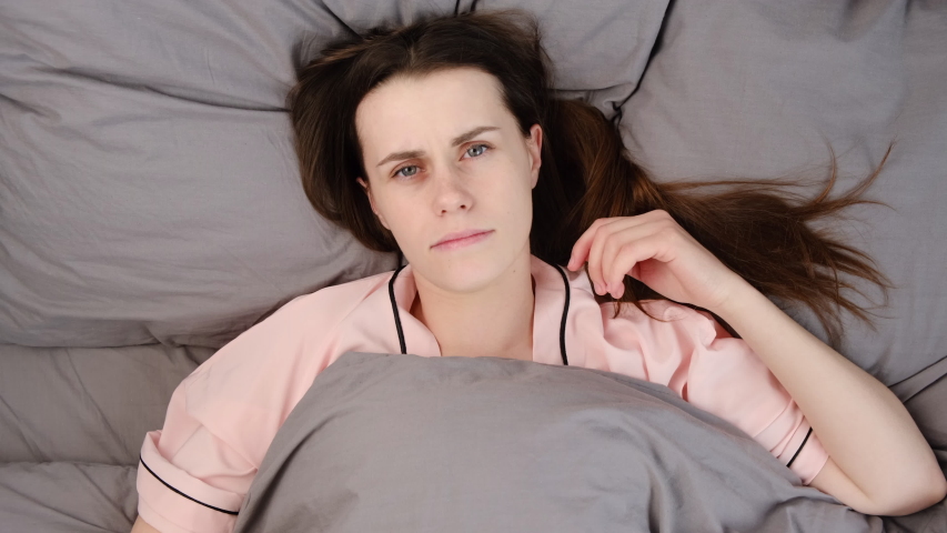Top view of discontent young woman has gloomy face,  has insomnia, cant get asleep late, lies under grey blanket, feels lonely.  Lady stressed because of too early wakeup, suffering from lack of sleep Royalty-Free Stock Footage #1050644752