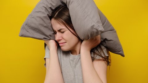 Young irritated woman trying to sleep, annoyed by bad noisy,  covering ears with pillow, angry girl student disturbed with loud noise problem,  suffer from insomnia, isolated on yellow background