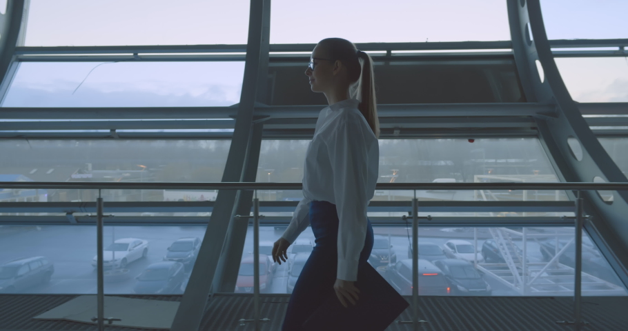 Side view of confident business lady walking in office hall against large windows. Female secretary holding clipboard of folder and going in corridor of business center on corporate meeting