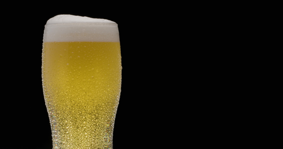 Glass of light beer on a black background. A jet slowly fills the glass with beer, causing abundant bubbles and foam. Clockwise rotation.