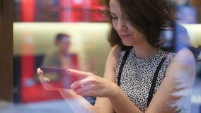 Woman watch video using smartphone, sit at cafeteria, shoot through window glass. Portrait shot with blurred background. She hold smartphone in hands, eyes fixed to display