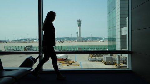 Silhouette of passenger woman walking against lounge window with trolley case, slow motion shot. Modern international airport, apron and traffic control tower seen outdoor