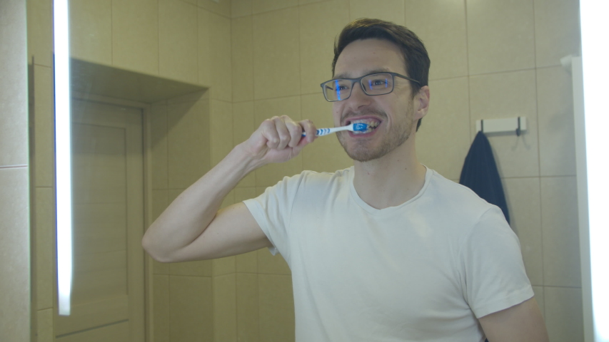 a man with glasses brushes his teeth in front of a mirror