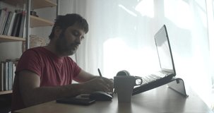 Man writing in a notebook on a wooden table with his video equipment and laptop. Work at home.