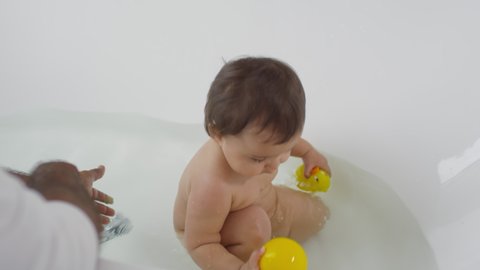 High angle shot of adorable toddler girl sitting in bathtub and holding yellow rubber toys, then laughing and showing ball to camera. Unrecognizable father watching over baby in bathtub