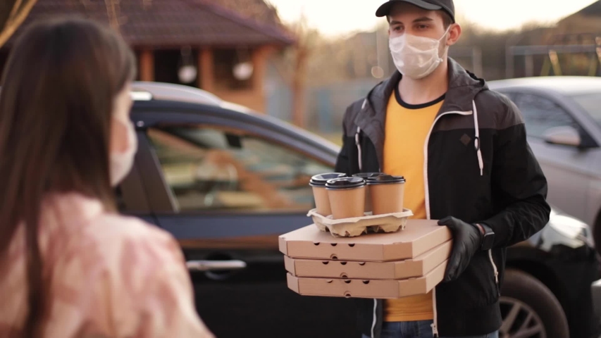 Young woman in protective mask and medical mask take order from delivery boy outside. Food delivery in quarantine. Courier bring Pizza and coffee for customer Royalty-Free Stock Footage #1050656941