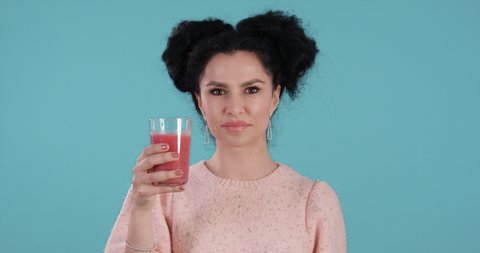 Portrait of beautiful young woman with black curly hair presenting and drinking red smoothie enjoyable in front of blue background shot in 4k super slow motion