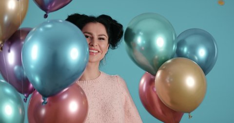 Portrait of beautiful young woman with black curly hair smiling dancing with ballons in her hands raining confetti in front of blue background shot in 4k super slow motion