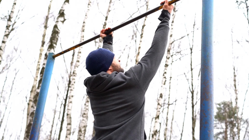 Close-up of a Mature man is outdoors and pulling up on a horizontal bar, he is wearing a warm sports uniform, a hat and special gloves for training.