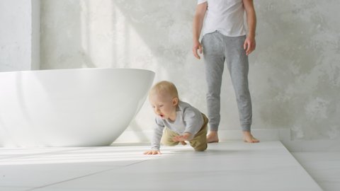 Medium shot of adorable baby girl with blond hair crawling on bathroom floor and giggling. Unrecognizable father standing in background