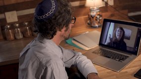 Jewish man with a beard in his 40s or 50s wearing a yarmulke holding a video chat with a female coworker, employee, or employer while working at home