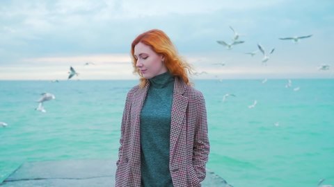 one redhead girl. stands on the pier by the sea, on the coast, by the ocean. winter or autumn, cold weather, birds and seagulls flying. wearing a coat. slow motion. lonely woman
