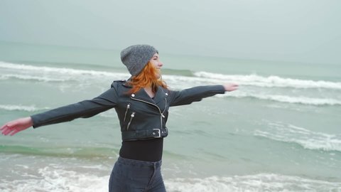 young girl in black clothes and leather jacket.  runs, whirls laughs at the camera by the sea or ocean.  winter or autumn, cold weather, slow motion, against the background of sand waves.  happy