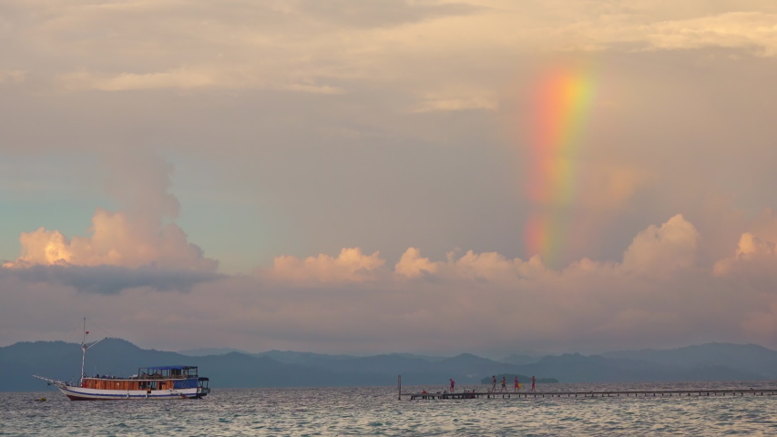Indonesia. Quiet evening over the tropical bay. Wooden jetty and pleasure boat. Part of the rainbow on a cloudy sky