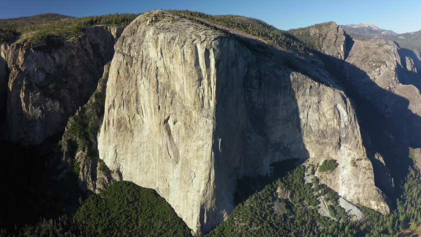 The flat vertical wall of the famous Mount El Capitan illuminated with early evening sunlight. Huge granite monolith of Sierra Nevada Range. Yosemite National Park. Aerial footage, 4K