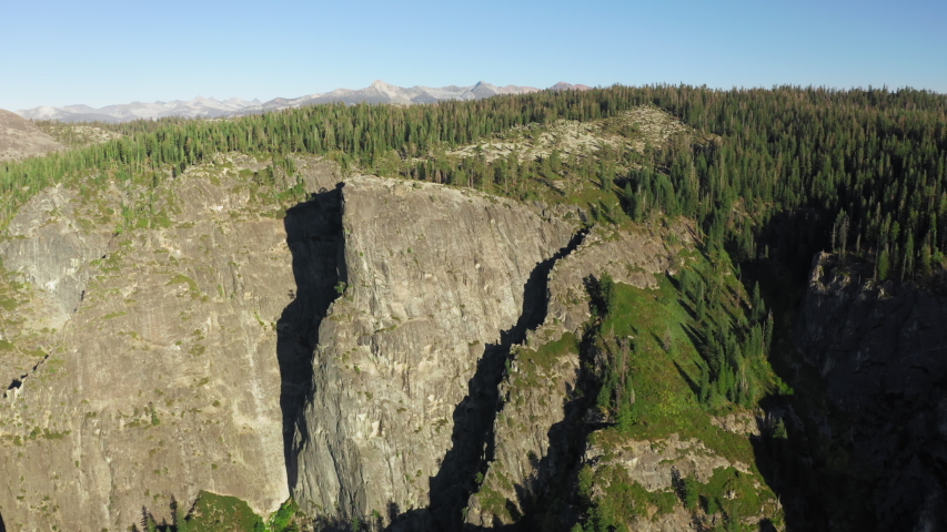 Aerial shot of large granite formations densely covered with conifers under clear blue sky. The top of the mountain is lit with the sun. Yosemite National Park, California, USA. 4K