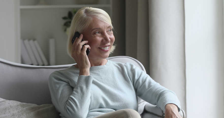 Happy middle aged woman relaxing on cozy sofa, talking on smartphone with friends. Excited elderly lady chatting sharing news on mobile phone with grown up children, resting in living room alone. | Shutterstock HD Video #1050697258