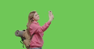 Young woman hiker with backpack walking isolated on green screen background at studio . Sporty girl dressed in pink hoodie making selfie portrait over chroma key. 4k raw footage video side view