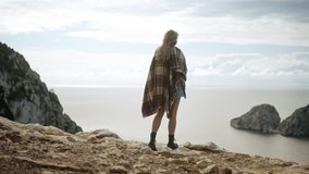 A young blonde woman walks on the cliff to the edge, in the background the island of Es Vedrà and the cloudy sky, Full HD long Shot Slow Motion Video