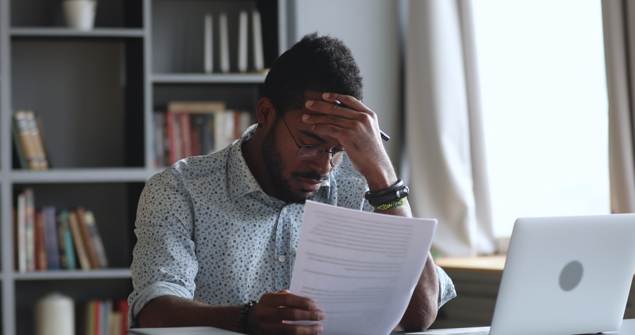 Thoughtful young african man looking through reading paper document, feeling stressed about banking loan money debt notification. Depressed biracial guy frustrated about high bills, dismissal notice. | Shutterstock HD Video #1050724879