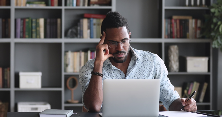 Thoughtful young african american man in eyeglasses watching educational lecture online on computer, writing notes on paper, web surfing searching additional information in internet, sitting at home. Royalty-Free Stock Footage #1050724882