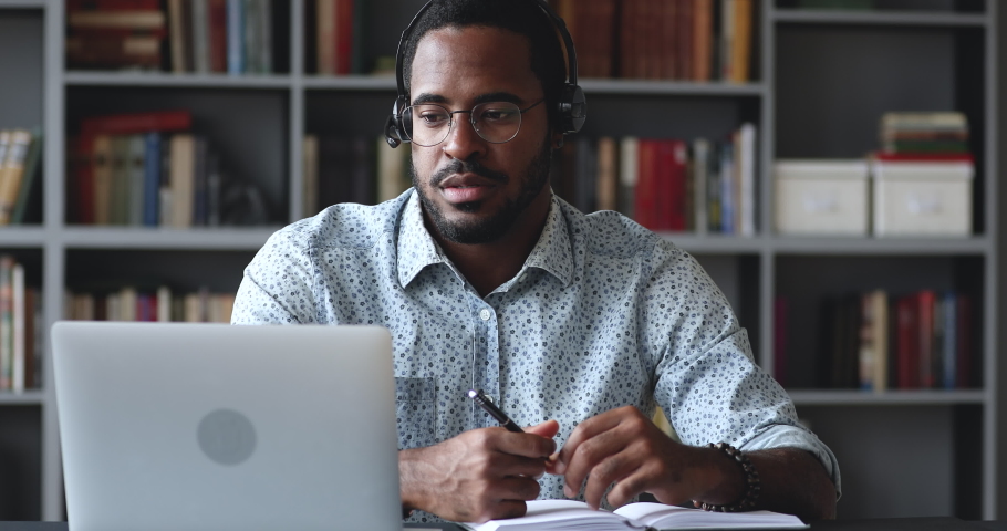 Happy young african american guy in glasses wearing wireless headphones with microphone, looking at laptop, holding video call talking speaking with consultant teacher mentor coach, writing notes. Royalty-Free Stock Footage #1050724894