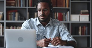 Happy young african american guy in glasses wearing wireless headphones with microphone, looking at laptop, holding video call talking speaking with consultant teacher mentor coach, writing notes.