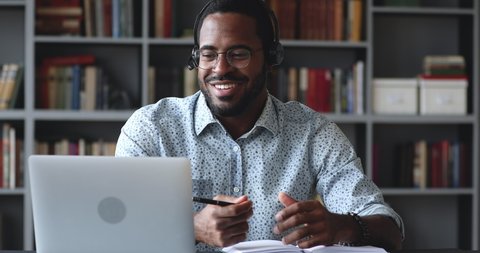 Happy young african american guy in glasses wearing wireless headphones with microphone, looking at laptop, holding video call talking speaking with consultant teacher mentor coach, writing notes.