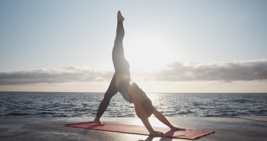The girl is doing fitness, frozen in a pose with a raised leg on a pier overlooking the sea.