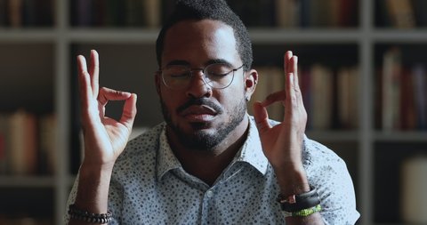 Close up head shot peaceful young african ethnicity man in eyewear making mudra gesture, reducing stress at work. Calm millennial mixed race guy doing yoga breathing exercises relaxing alone at home.