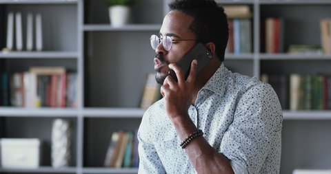 Serious young african ethnic man in glasses talking on smartphone with service operator, discussing solving problems by mobile phone. Confident mixed race businessman calling talking to client.