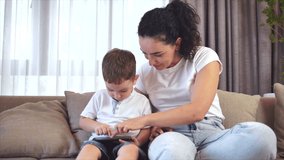 Cute family happy mom and cute little kid son look at screen of a tablet cell phone, mother with child sit on the couch at home playing on the smartphone laugh uses a modern application.