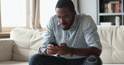Focused young biracial guy sitting on sofa, using mobile apps on phone, web surfing online, searching information. Serious african businessman reading email, typing message on smartphone indoors.