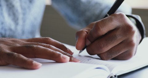 Close up young african american male student holding pen, writing notes in paper notepad. Confident mixed race businessman planning workday, noting daily cases in organizer before workday start.