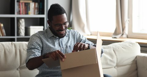 Curious happy young african american man unboxing large carboard parcel, feeling satisfied with fast delivery or purchases item. Excited male biracial client unpacking order from internet store.