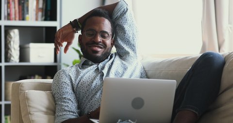 Happy young african american man in eyeglasses relaxing on cozy couch, looking at laptop screen, chatting with friends in social networks, dating online, enjoying leisure weekend time alone at home.