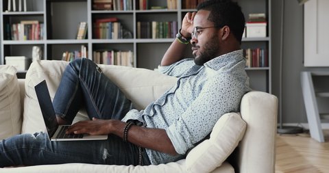 Side view young smart multiracial guy in eyeglasses leaning on comfortable sofa, working remotely on computer from home. Focused millennial african ethnic student studying distantly on online courses.