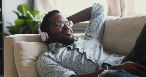 Happy millennial biracial hipster guy in glasses wearing modern wireless headphones, choosing enjoying favorite music from mobile application, relaxing alone on comfortable couch in living room.