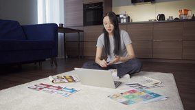 Young Asian woman sitting cross-legged on floor at home and spraying antiseptic sanitizer on her hands while having video conference with colleagues on laptop 