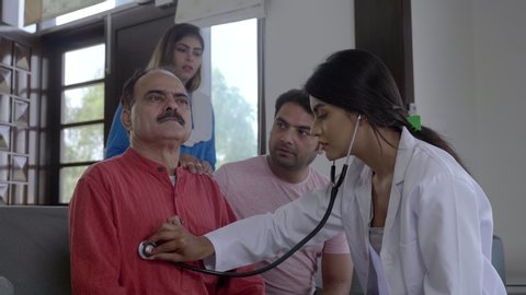 A wide shot of a doctor interacting with concerned family members about health of an old patient. A friendly female physician talking checking examining grandfather senior elder man in the house