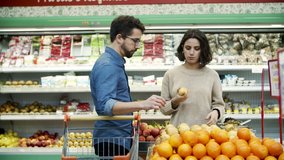 Couple buying fruits in supermarket. Focused young man and woman standing with shopping trolley and choosing fresh pears and oranges in grocery store, handheld shot. Shopping concept 