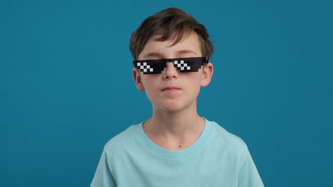 Portrait happy boy chewing gum and learning to inflate bubbles and bursting in pixel sunglasses it joyfully smiling looking at camera slow motion on blue background. Childhood. Emotions people