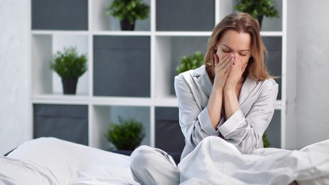Sick girl in sleepwear cough and sneeze sitting on bed covering face by napkin. Morning lady waking up at bedroom feeling influenza symptoms. Shot with RED camera in 4K