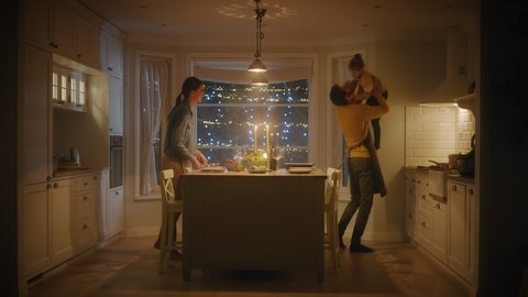 Happy Family of Three Cooking and Having Dinner Together. Mother Prepares and Serves Food, Cute Little Girl Runs to Father, Hugs Him and They Dance. Festive Table in Cozy Kitchen Interior