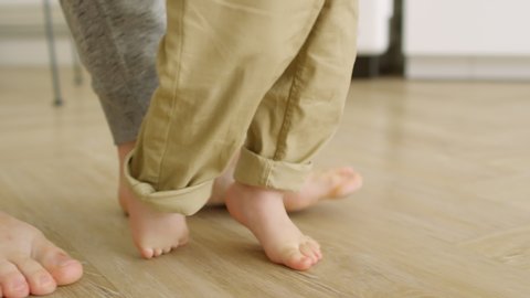 Tracking with side view of unrecognizable father supporting baby making first steps on floor – Stockvideo