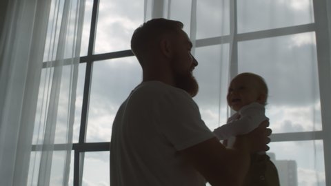 Medium shot with backlit of bearded father holding cute baby and twirling by window on gloomy day స్టాక్ వీడియో