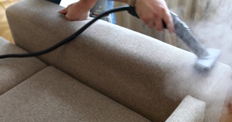 Male housekeeper hand cleaning sofa with steaming brush closeup. Professional house wash business concept