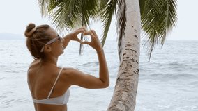 Young woman making heart shape finger frame on idyllic wild beach. People travel vacations love concept. Girl sitting on palm tree loving nature and ocean 