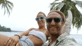 Young couple taking selfie on tropical beach, woman sitting on palm tree waving hello to camera while video chatting online sharing adventure and vacations with friends. Couple selfie travel