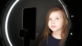 Small girl using mobile phone, speaking at camera of smartphone in front of ring light, shooting video for blog. Adorable child learning new technology. 